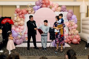 People perform on a stage decorated with pink and purple balloon arch during an indoor event.