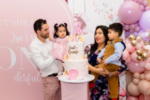 A family celebrates at a pink-themed first birthday party with a decorative cake and balloon arch in the background.