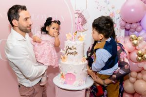 Family gathered around a white and pink unicorn birthday cake with balloon decorations at a pink-themed party celebration.