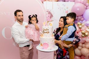 Family poses together at pink balloon-decorated party with cake and festive decorations.