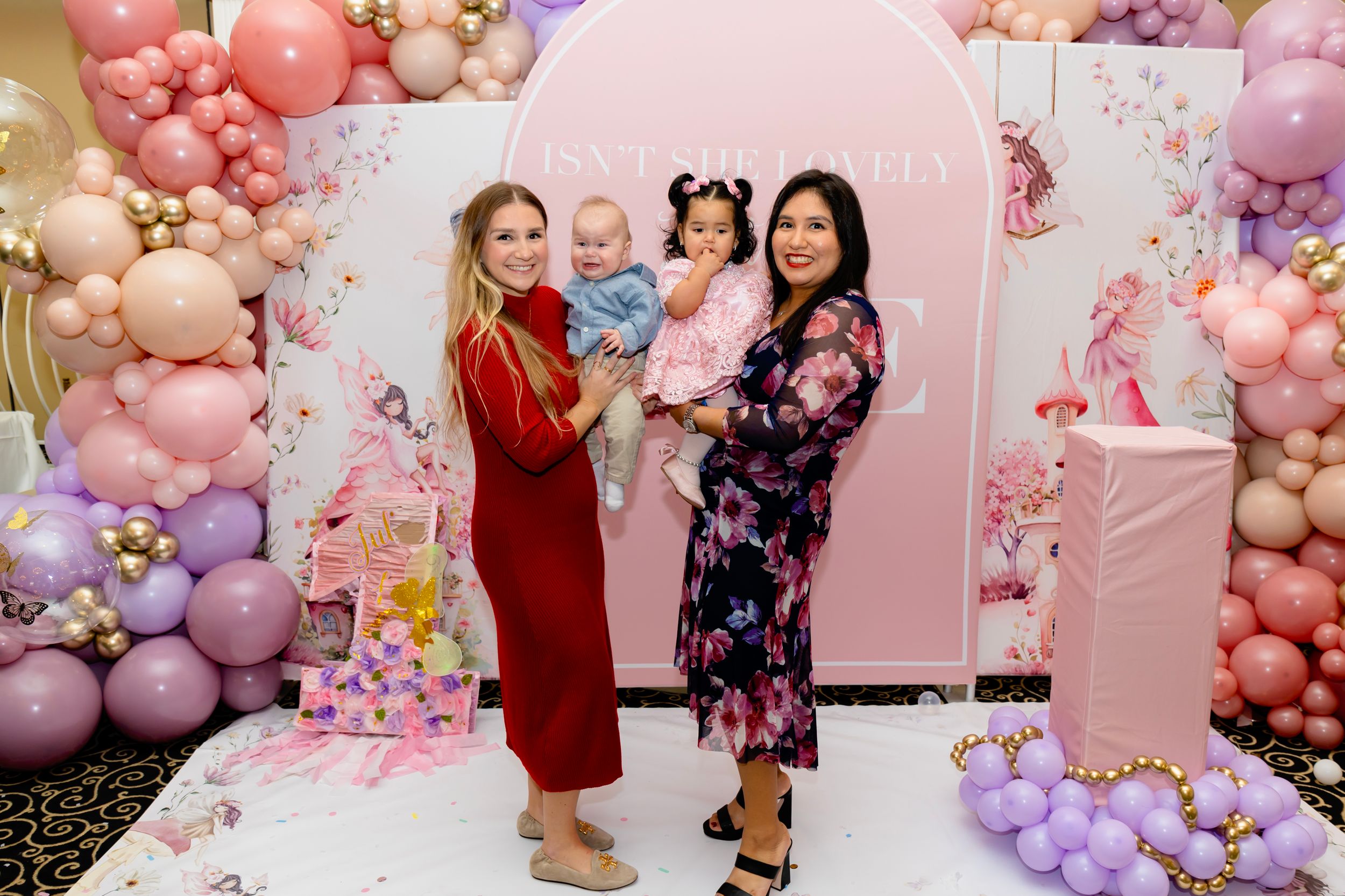 Elegant party setup with pink and purple balloon garlands and floral backdrops at an indoor celebration.