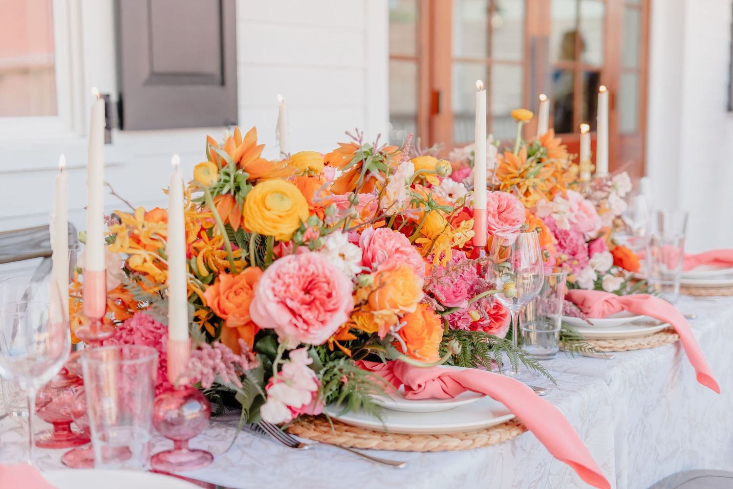 Elegant table setting with vibrant coral and orange floral centerpiece, pink ribbons, and tall white candles on white linens.