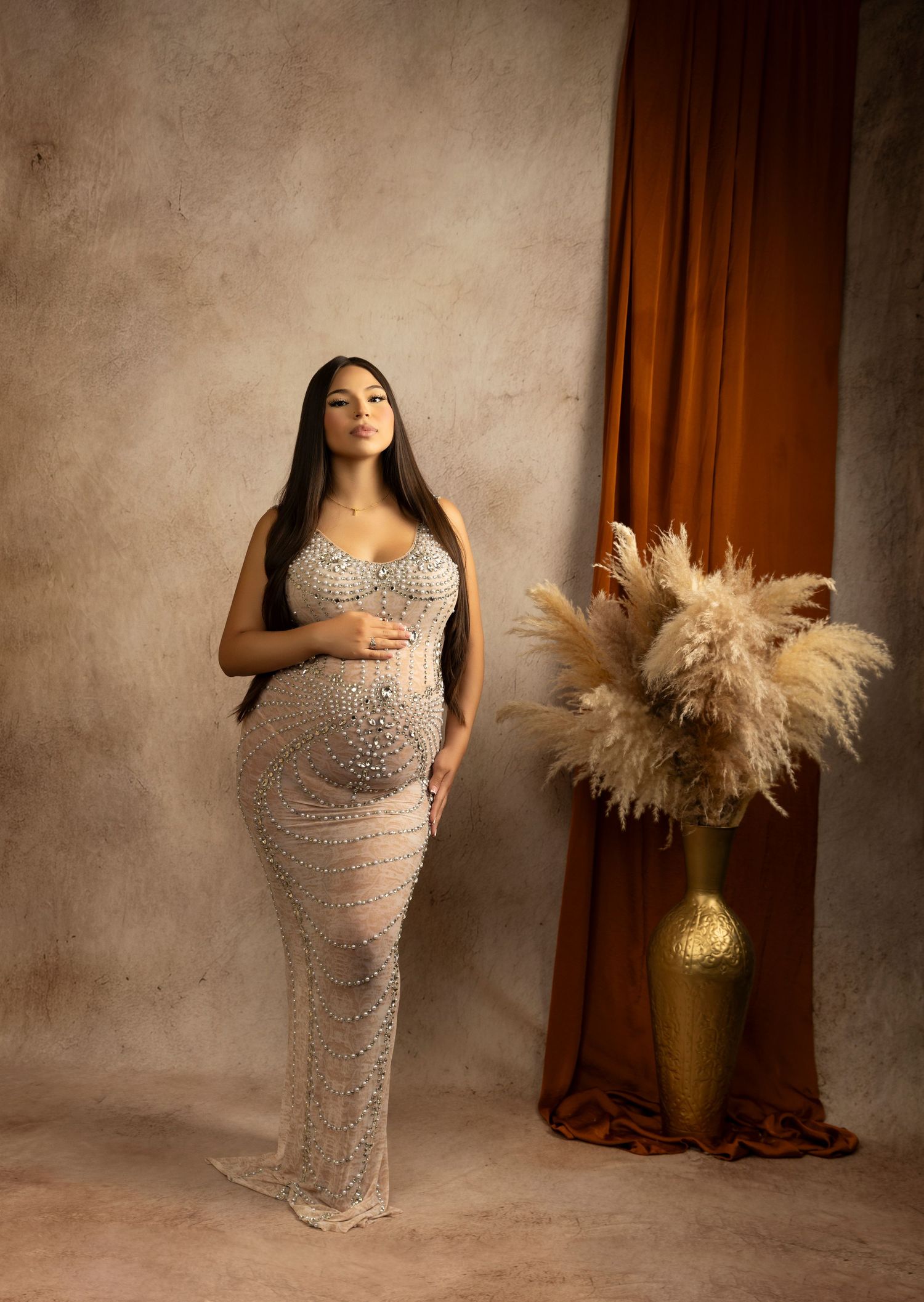 Woman in sparkling silver maternity gown poses beside gold vase with pampas grass.