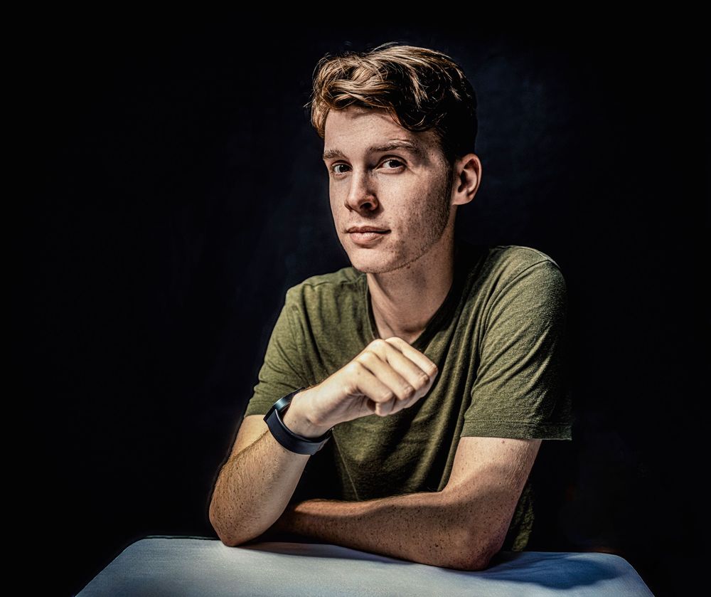 Subject in olive green t-shirt poses with arm resting on blue surface against dark background in dramatic portrait lighting.