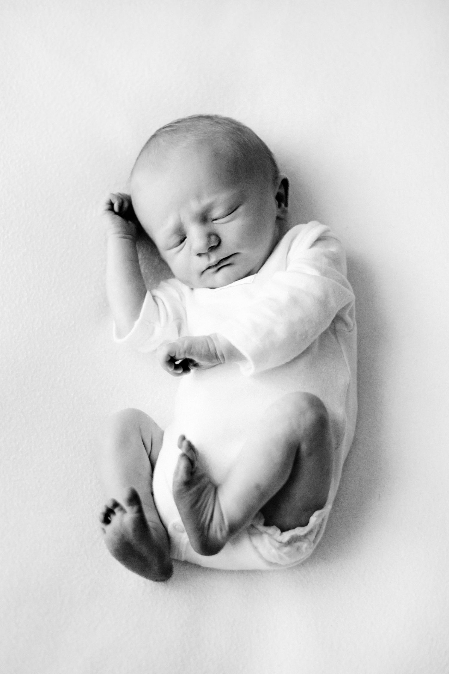 A peaceful sleeping newborn baby in white clothing lies curled up on a light background in black and white photography.