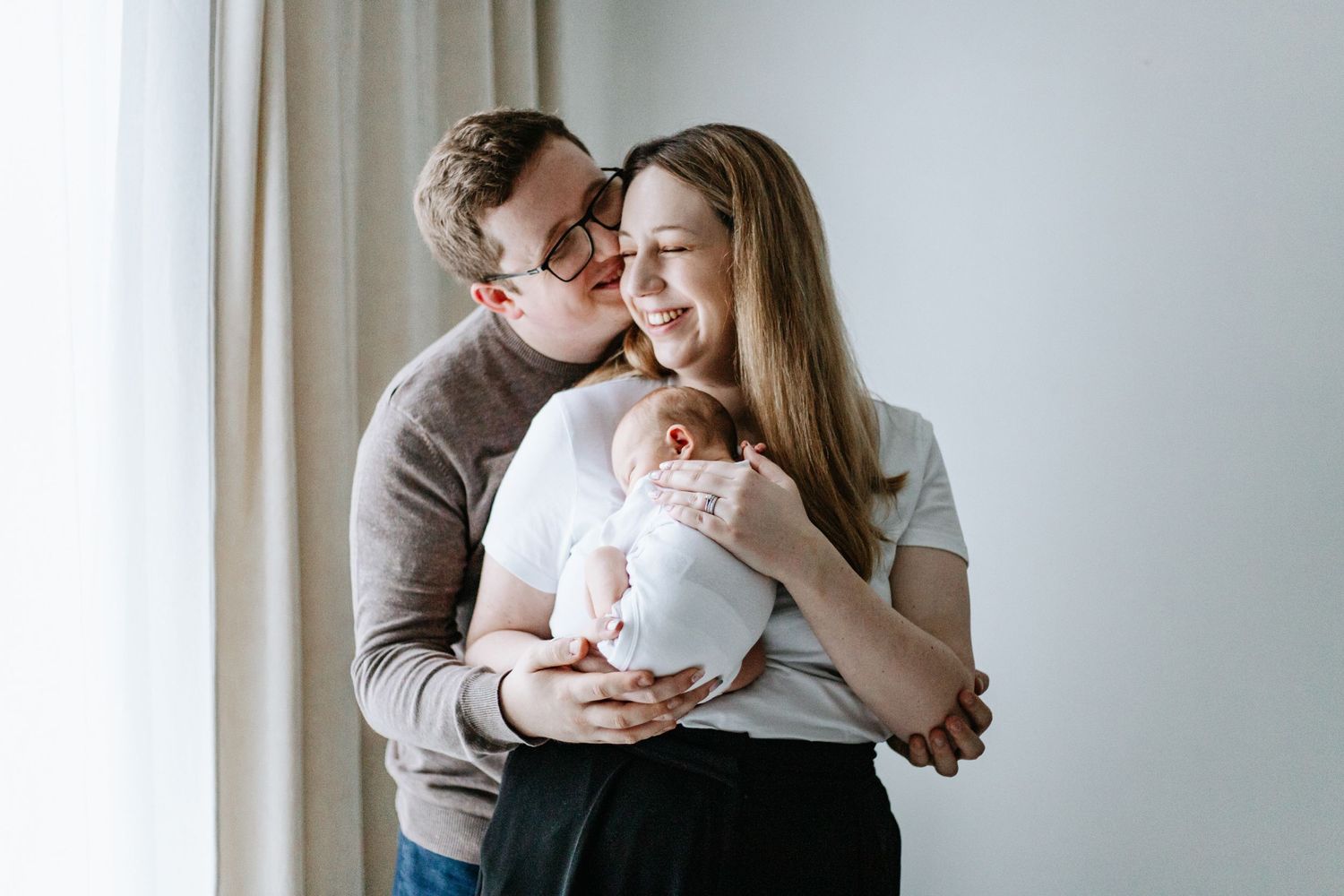 A tender family moment as parents embrace while holding their newborn baby beside a bright window.