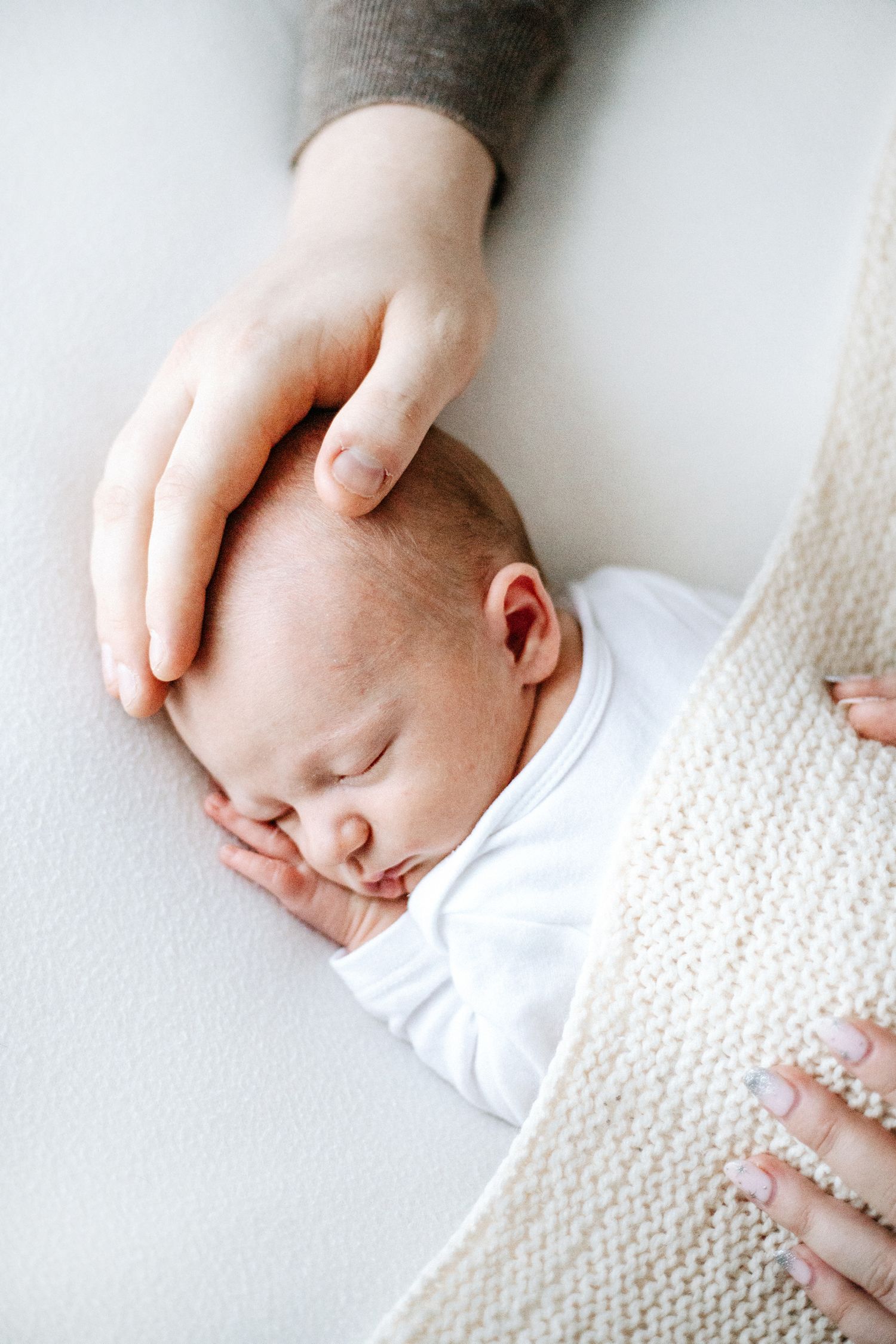 A sleeping newborn baby in white swaddled in a knit blanket with a gentle hand resting on their head.