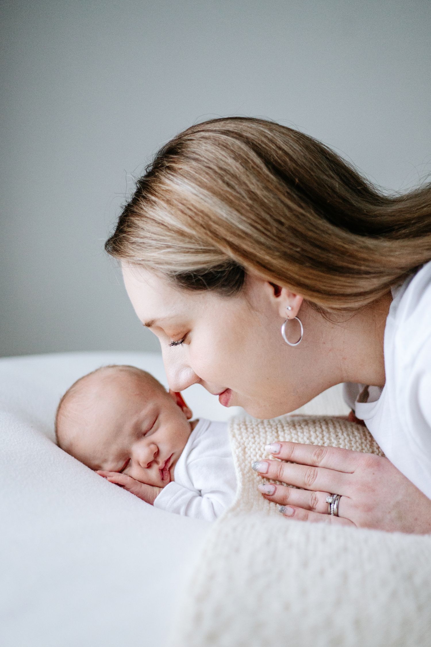 A tender moment between mother and sleeping newborn baby on white bedding, captured in soft natural light.