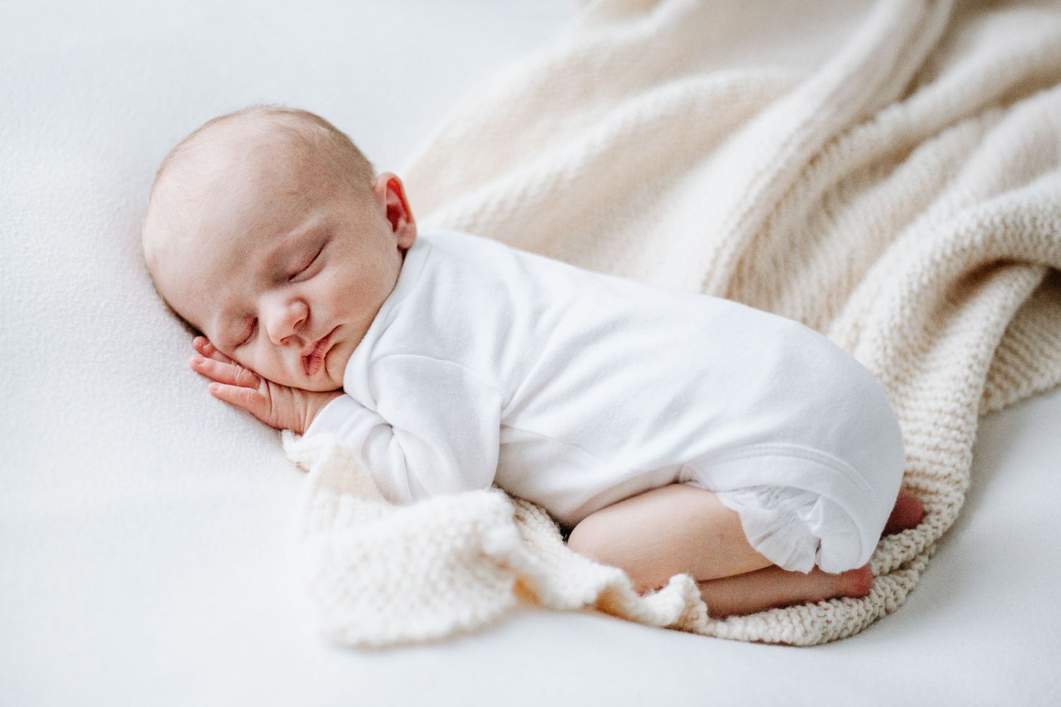 A peaceful sleeping baby in white clothing rests on a soft cream-colored knit blanket.