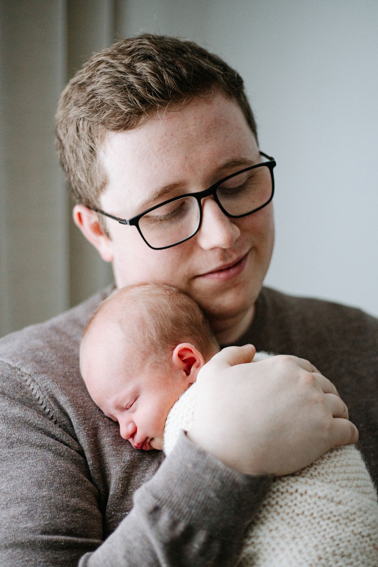 A person wearing glasses and a grey sweater tenderly holds a sleeping newborn baby wrapped in a white blanket.