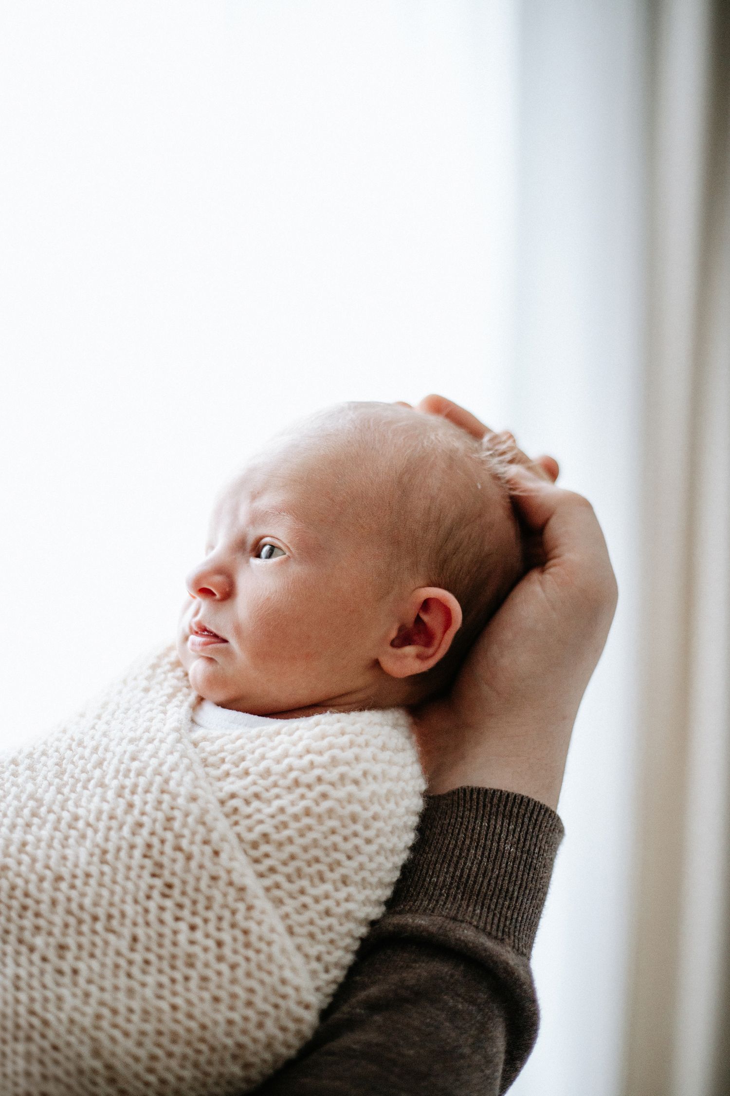 A small infant wrapped in a cream knit blanket is gently cradled by an adult hand near a bright window.