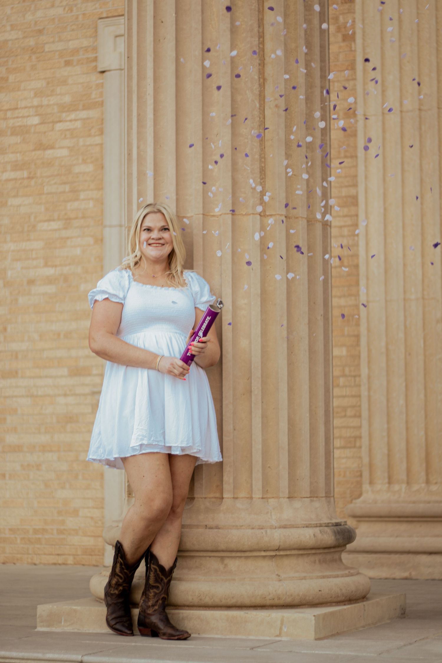 A graduate in a white dress and cowboy boots stands by columns while confetti falls during a celebration photoshoot.
