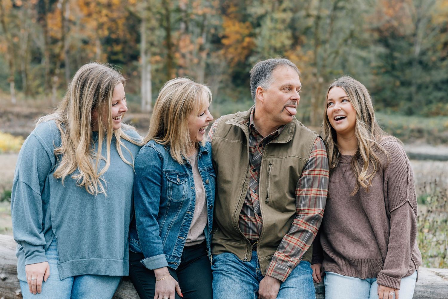 Two parents and two daughters laugh together outdoors surrounded by vibrant fall foliage.