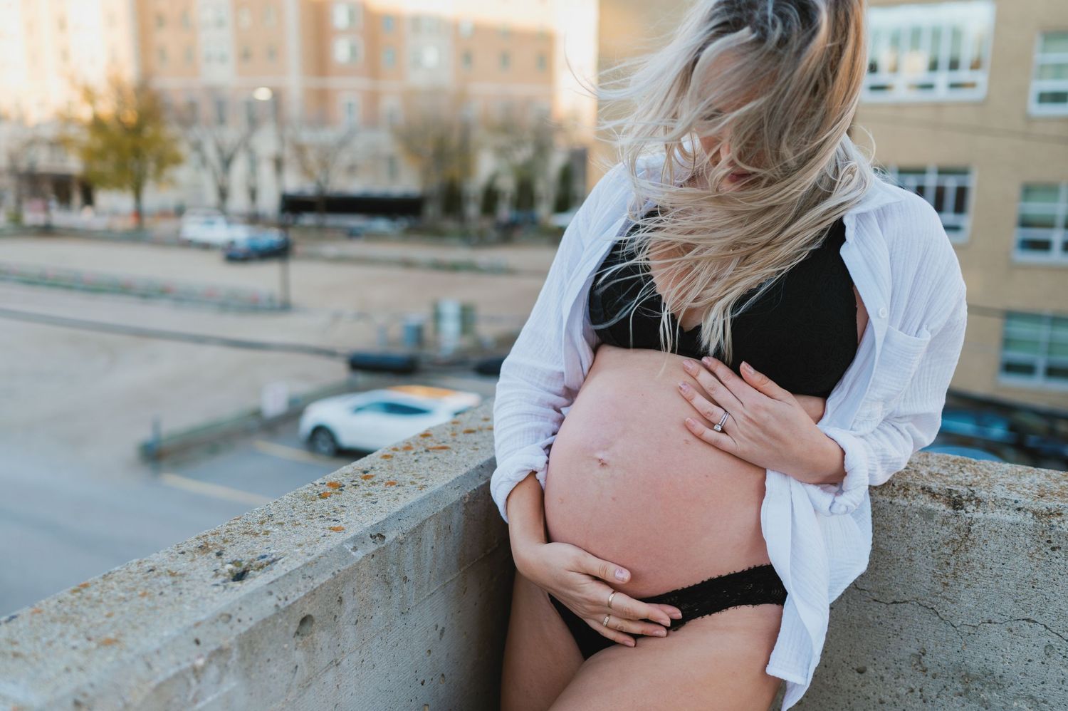 Expecting mother in black lingerie and white shirt cradles her baby bump on a city rooftop.