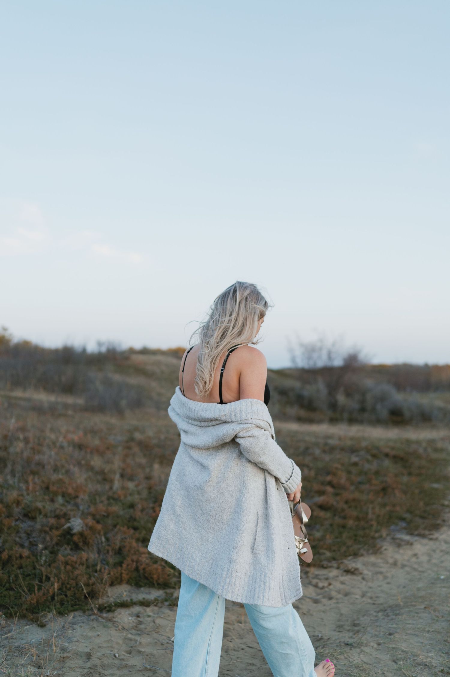 A figure in a cozy sweater and light pants walks along a scenic hillside path at dusk.