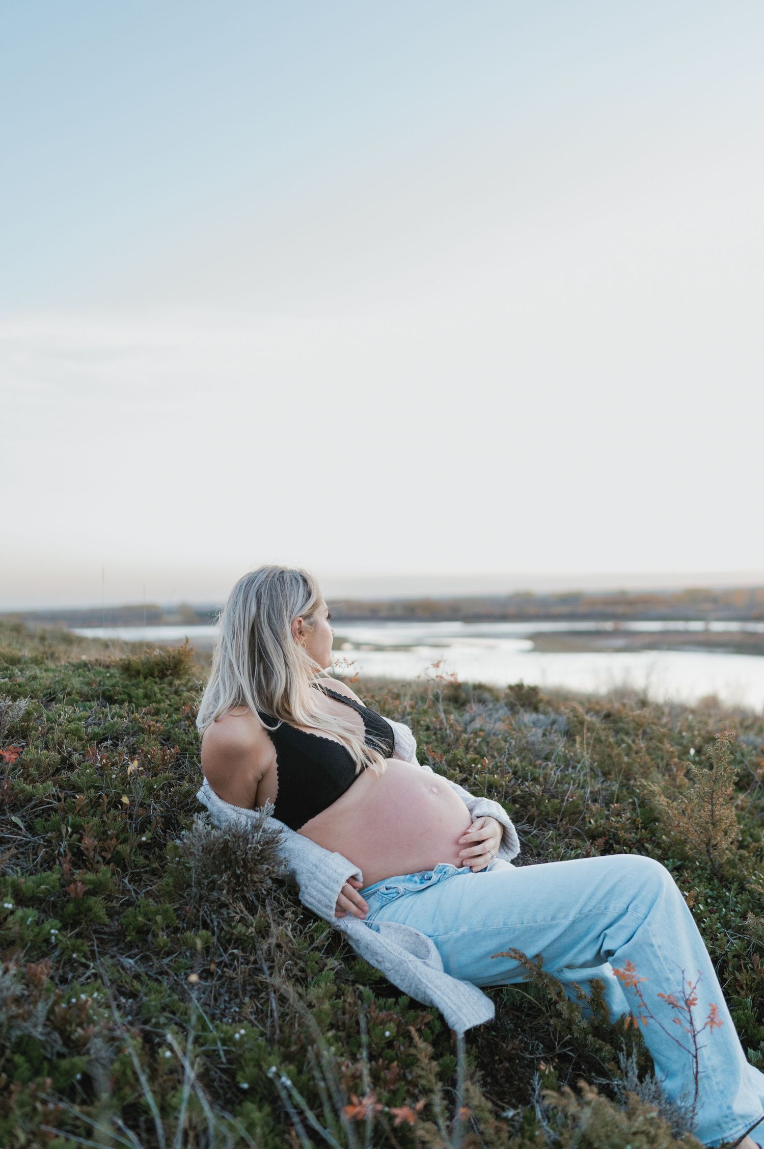 An expectant figure in a black top and light jeans rests on a grassy hillside at sunset.