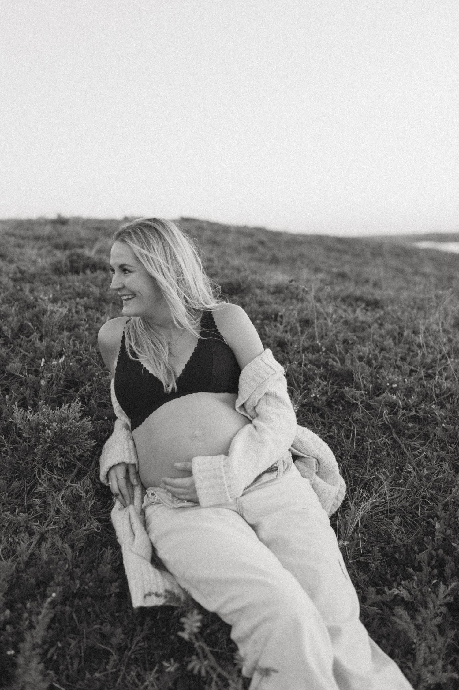 A black and white maternity photo captures a candid moment on a grassy hillside.
