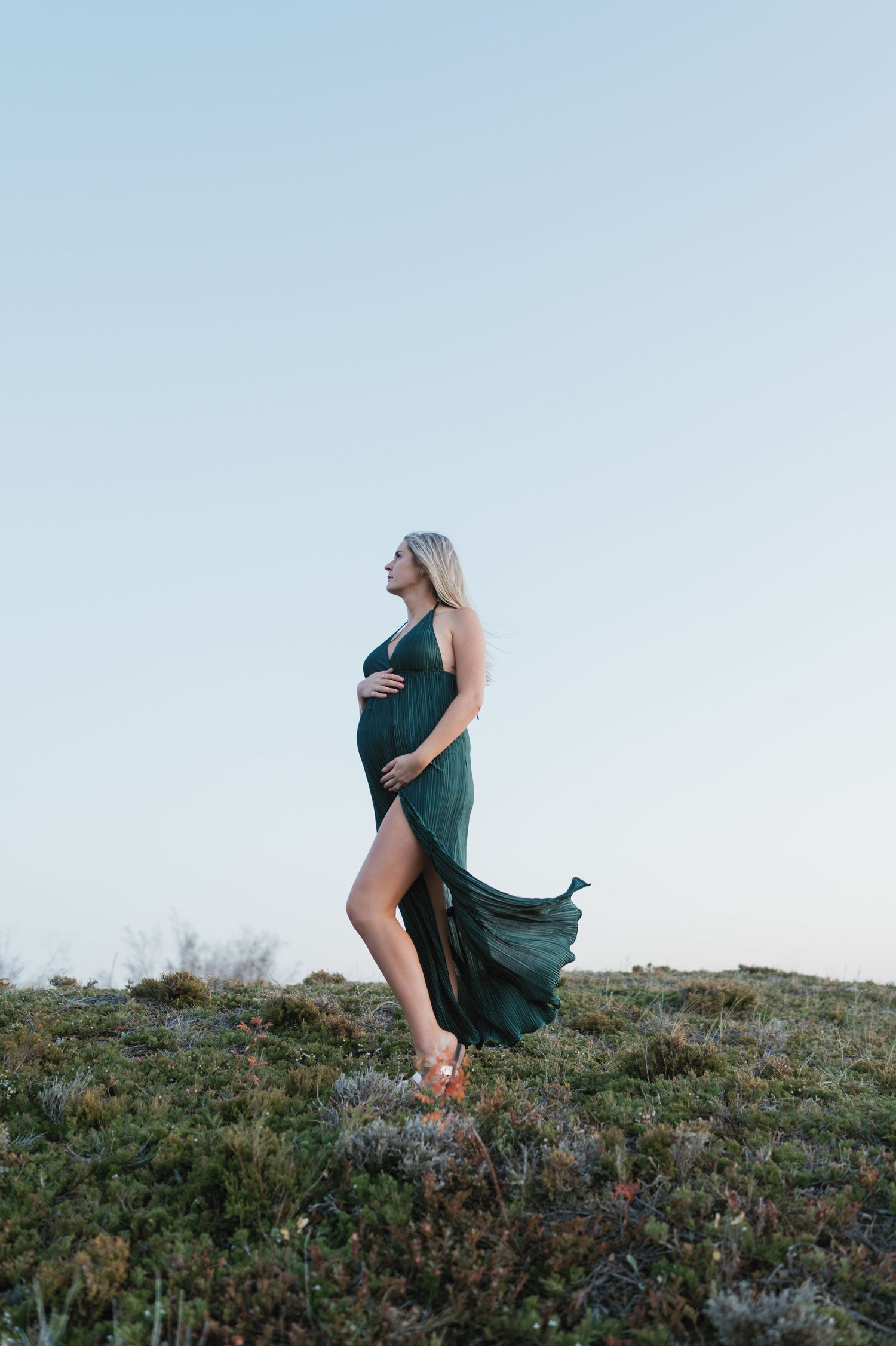 Expectant woman in flowing emerald dress stands on grassy hilltop against light blue sky.