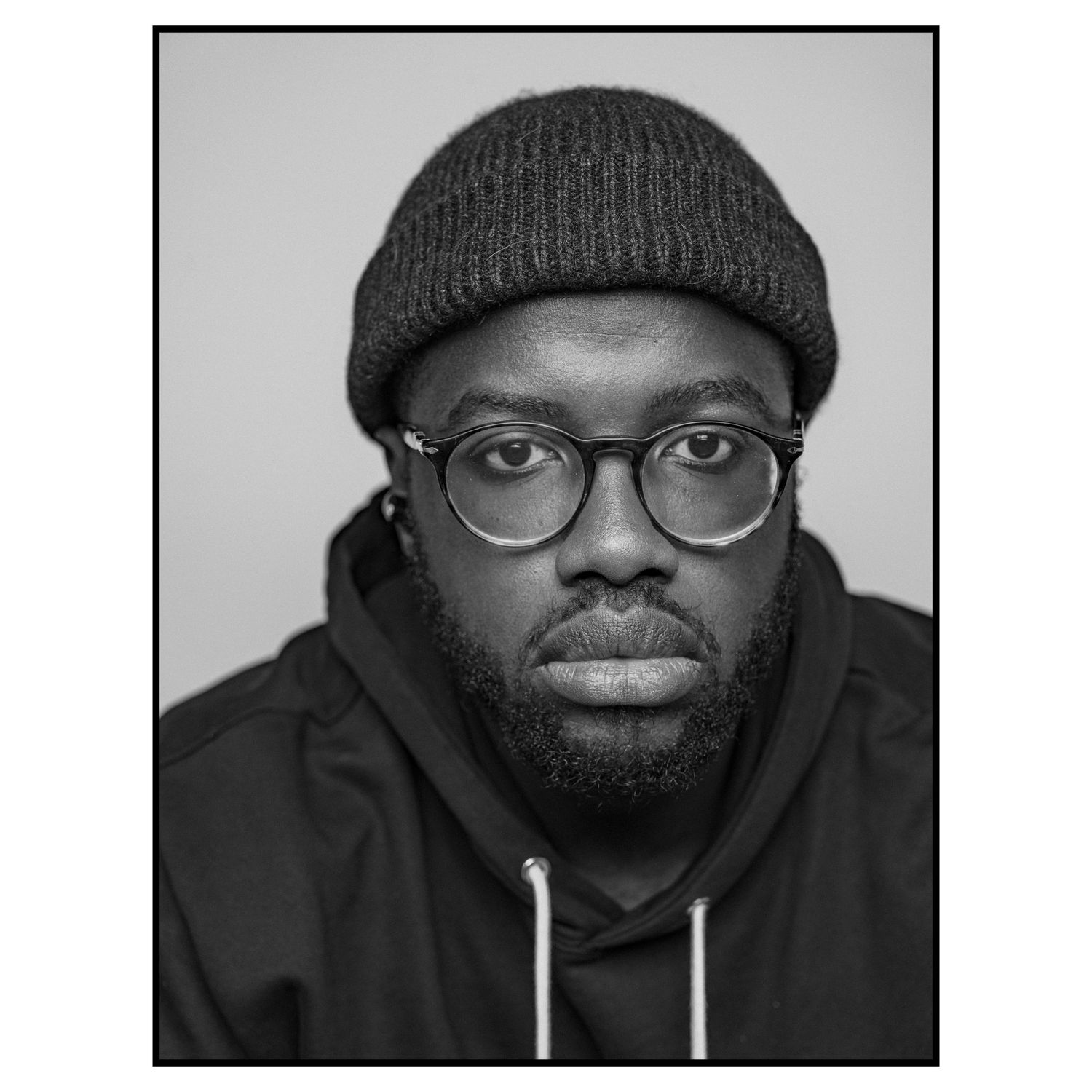 A portrait shot of a person wearing a beanie hat, glasses and hoodie against a gray background.