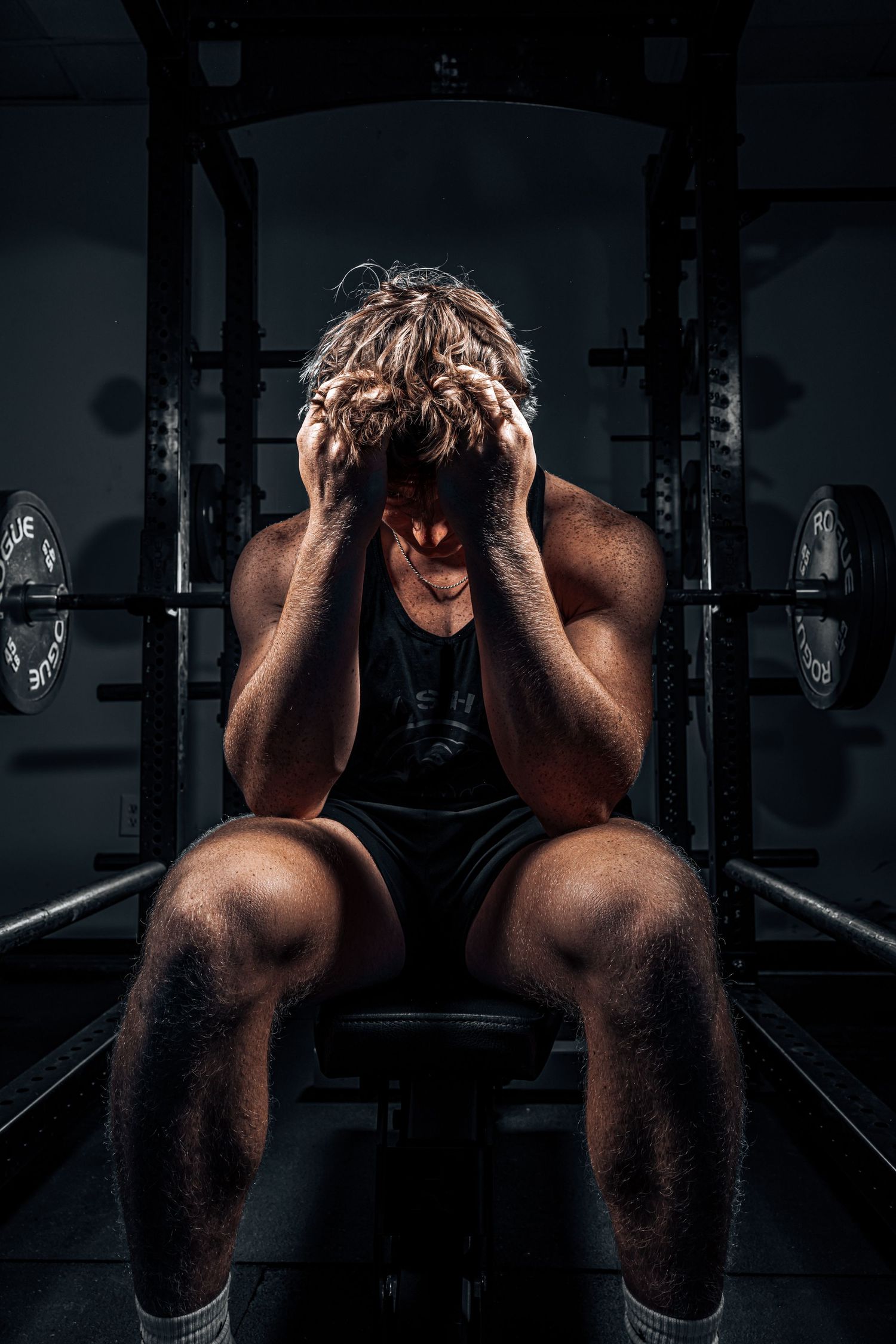 An athlete shows intense emotion while resting on a weight bench in a dark gym setting.