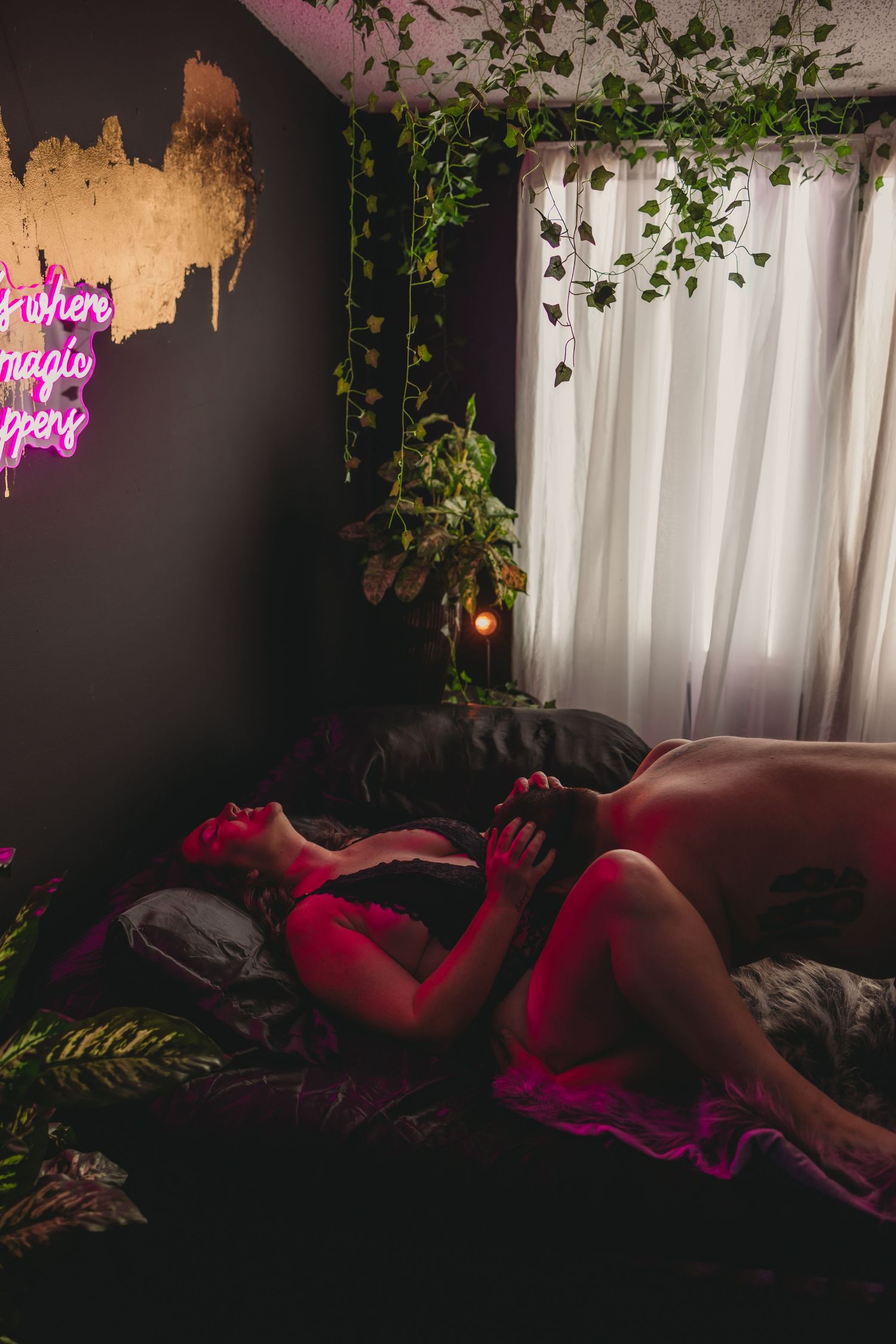 Moody room with pink neon sign, trailing plants and white curtains illuminated by red lighting.