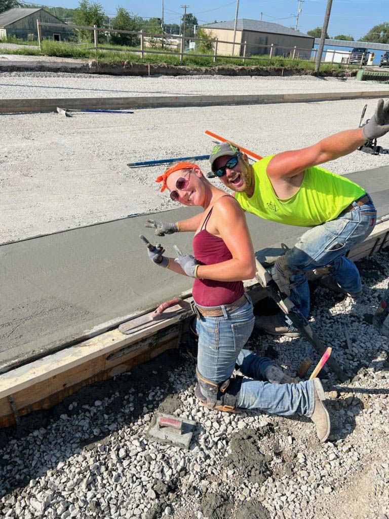 Construction worker in bright safety vest poses playfully while working on concrete foundation.