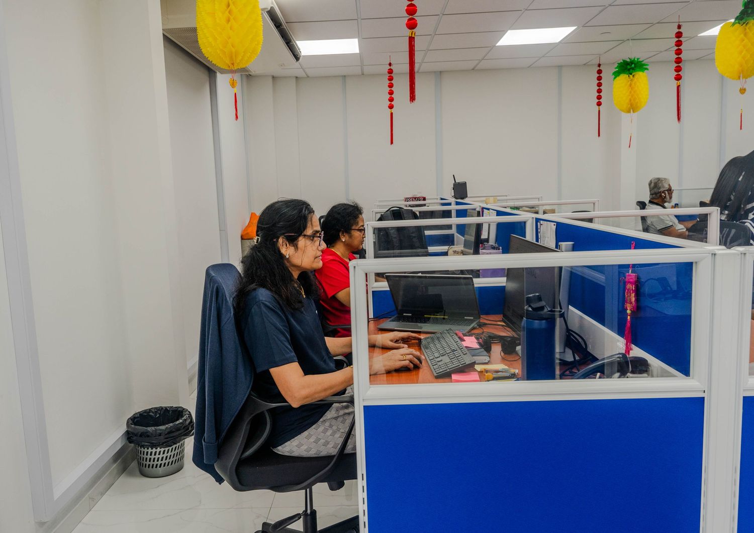 Office workers at blue cubicle workstations with festive decorations overhead.