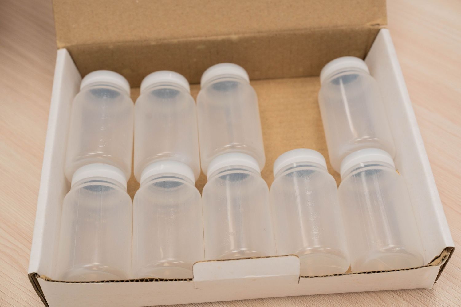 Box containing ten clear plastic laboratory bottles with white caps arranged in neat rows.
