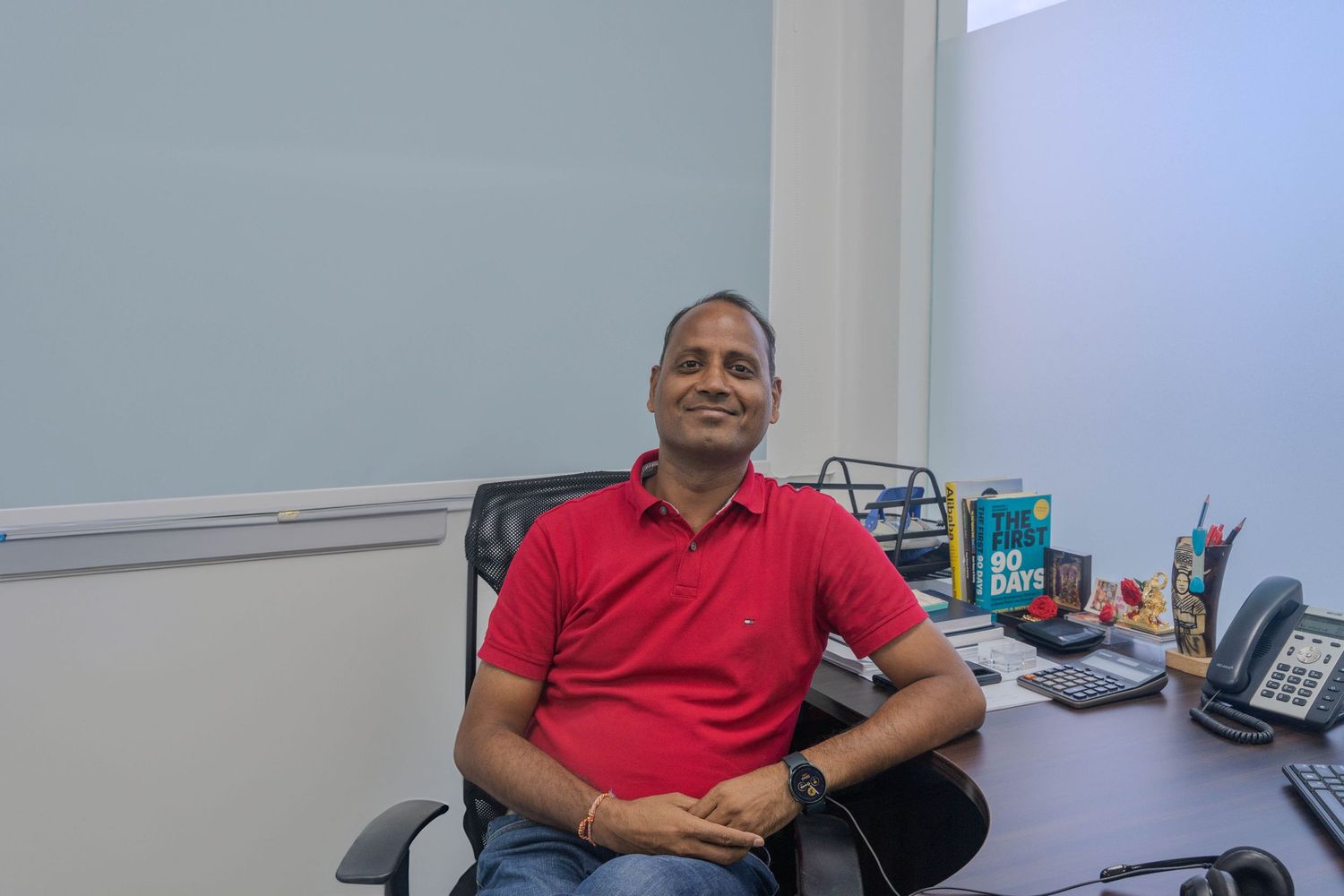 Employee in red polo shirt sitting at office desk with computer and supplies.