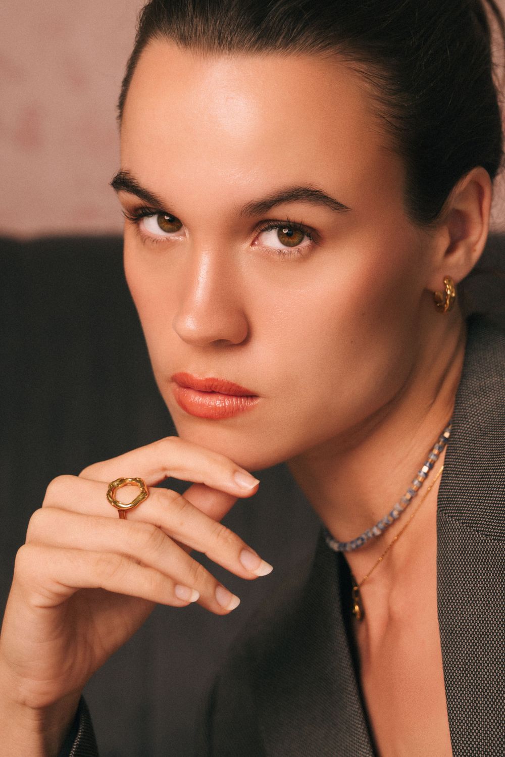 Studio portrait showcasing elegant jewelry and makeup with dark blazer against neutral background.