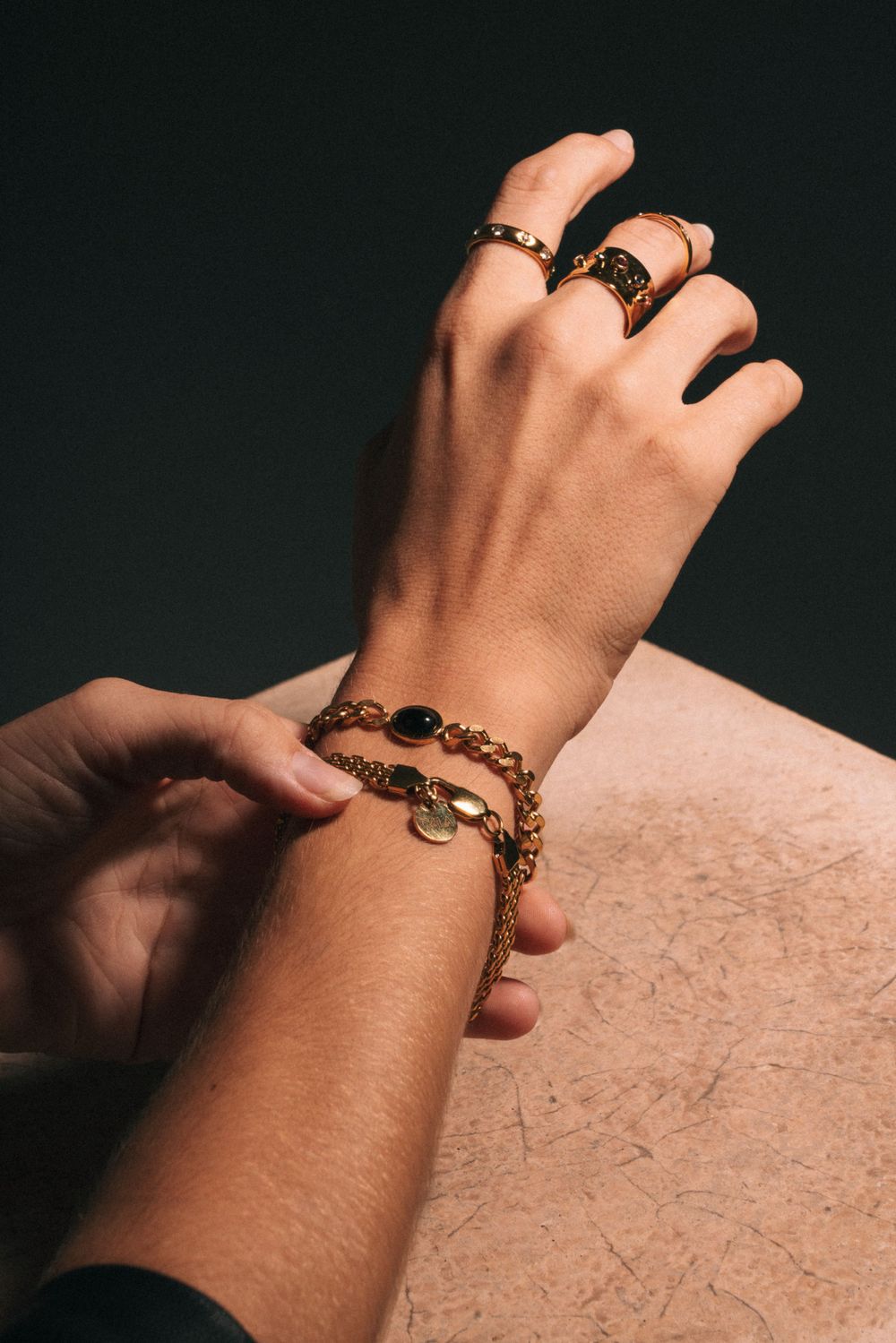 Close up of hand wearing multiple gold rings and chain bracelet against dark background.