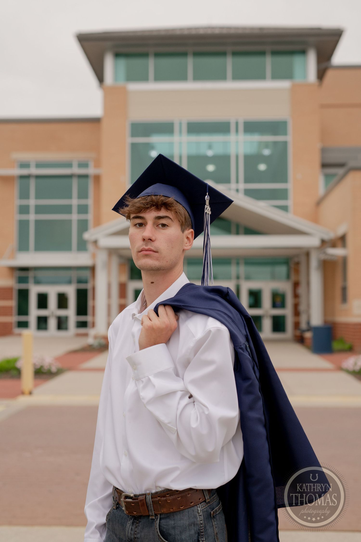 Graduate in cap holds gown over shoulder while standing in front of school entrance.