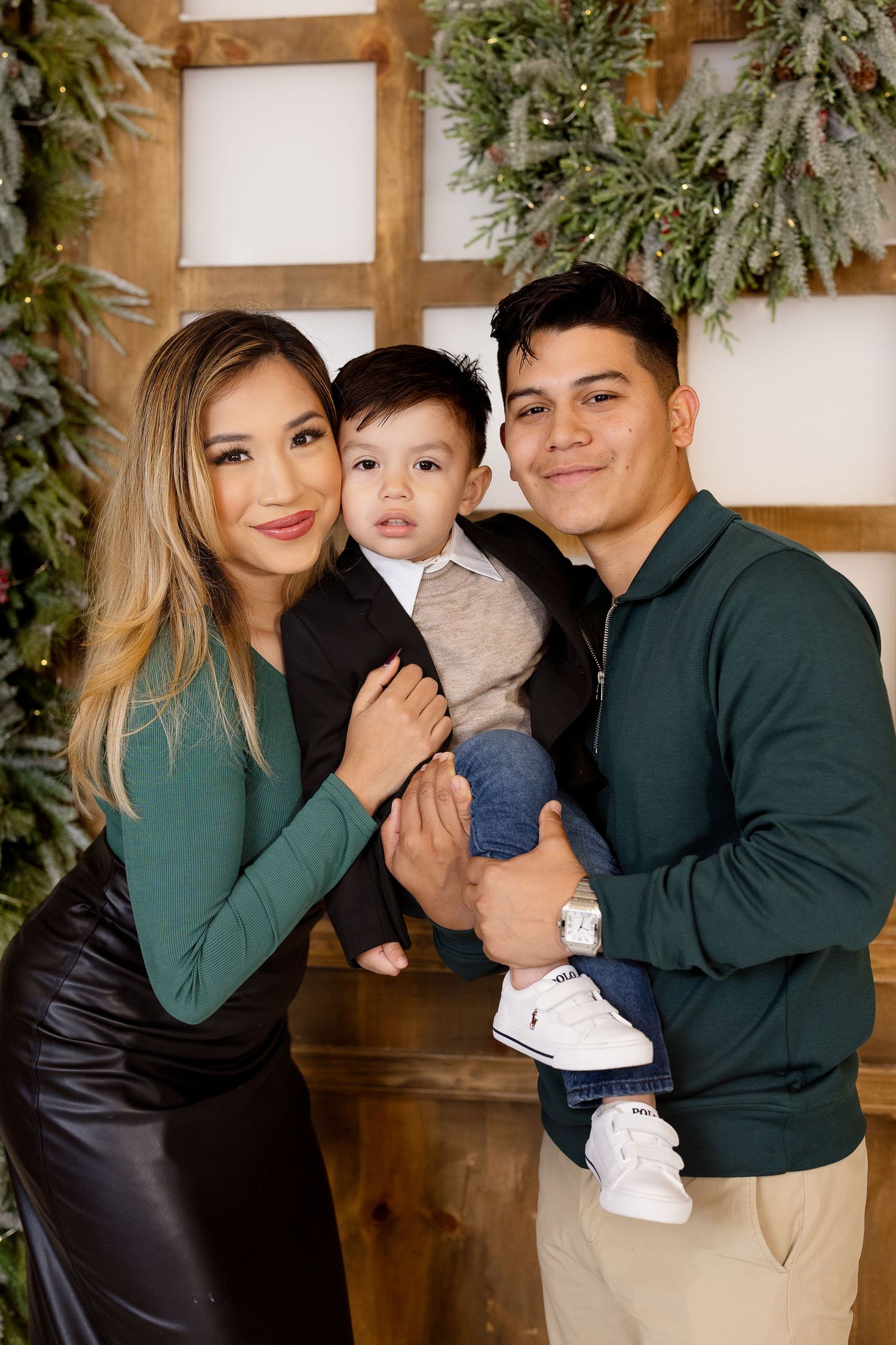 A family poses together in front of a wooden backdrop with greenery wearing coordinated green and neutral outfits.