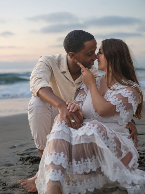 A romantic beach photoshoot featuring a couple in white attire sharing an intimate moment at sunset.
