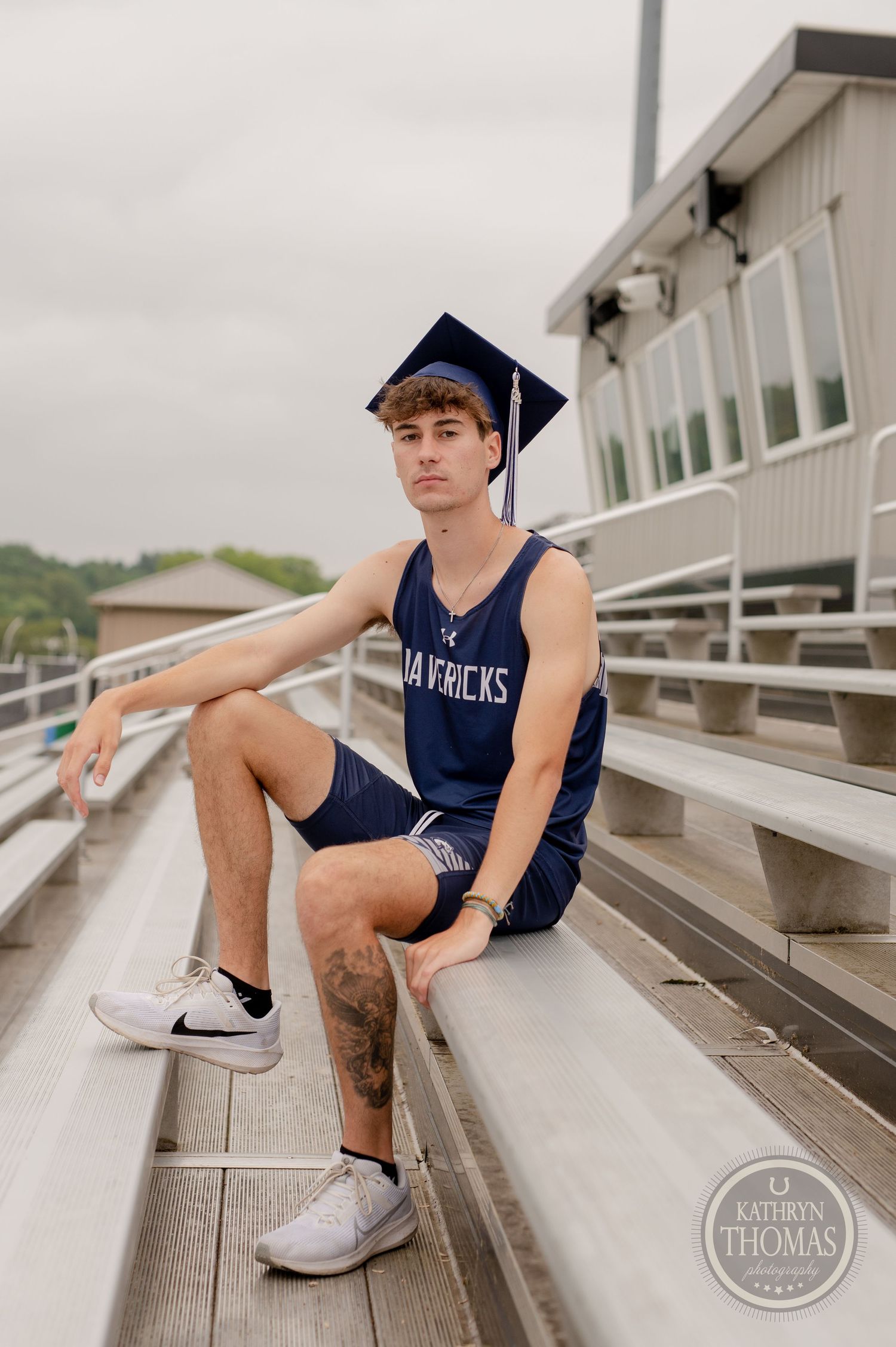 A graduate in navy blue athletic wear sits on metal bleachers on an overcast day.