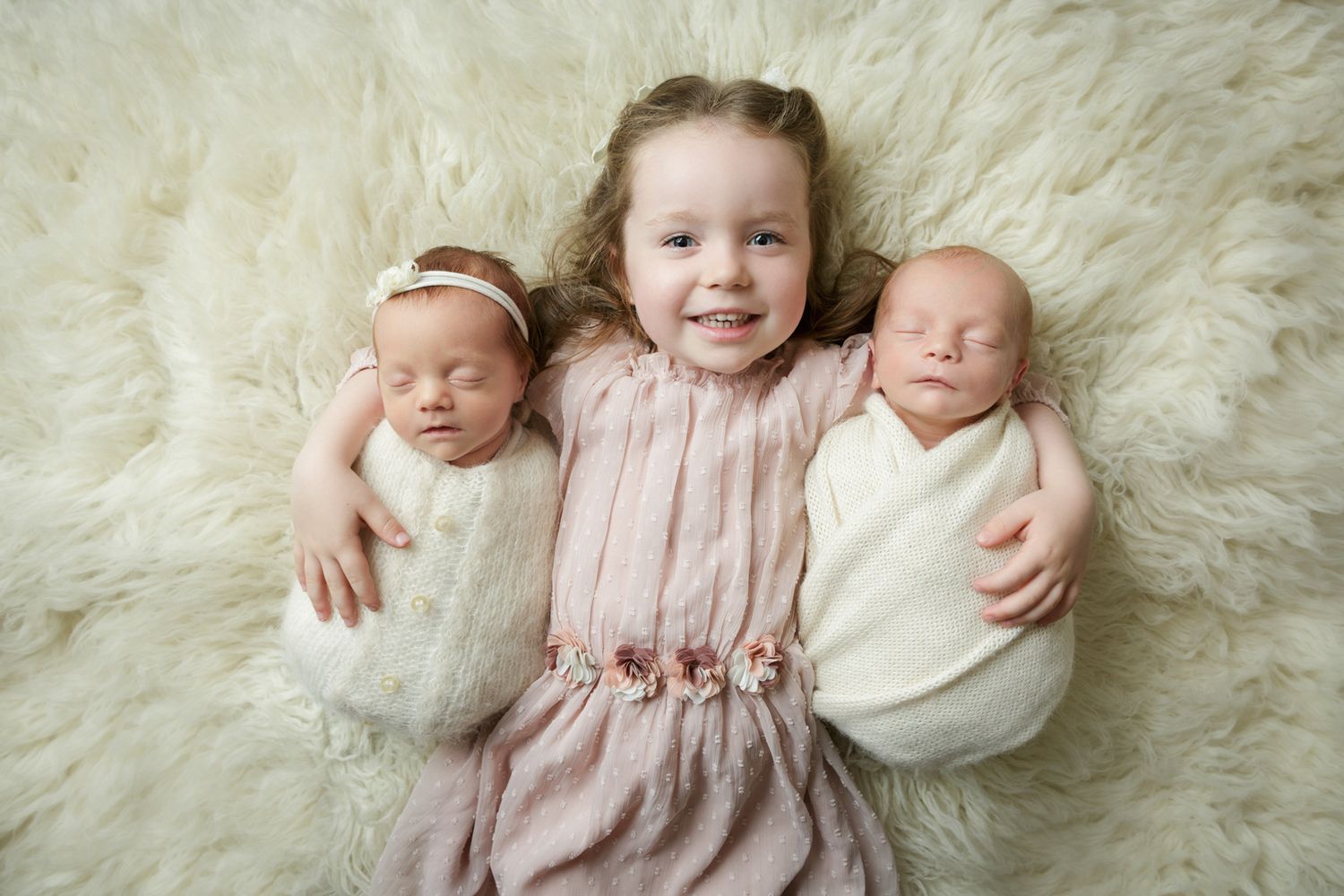 older sister lying on a fluffy blanket cuddling her twin baby siblings