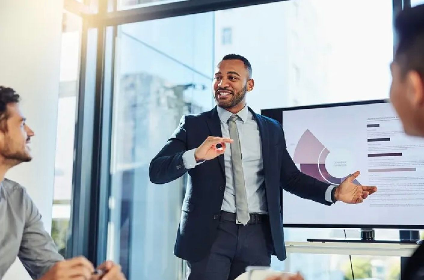 Business professional in a dark suit gives a presentation while gesturing near a conference room whiteboard.