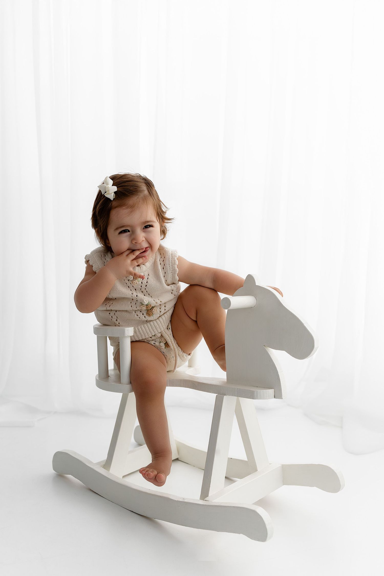 A small child sits happily on a white wooden rocking horse against a light background.