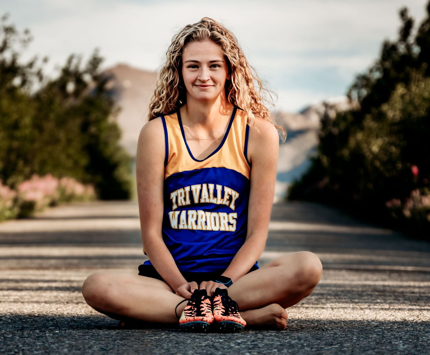 A high school athlete in blue Warriors uniform sits cross-legged on an outdoor road.