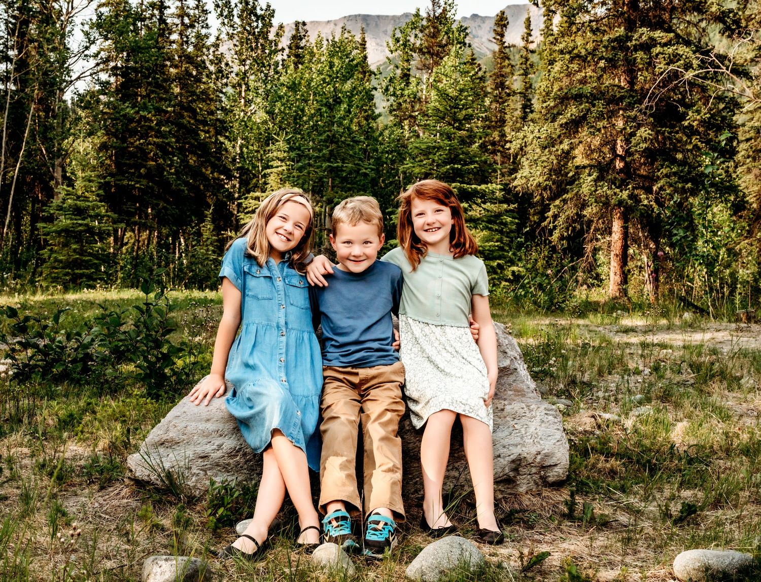 Three children sit together on a rock with mountains and pine trees in the background.