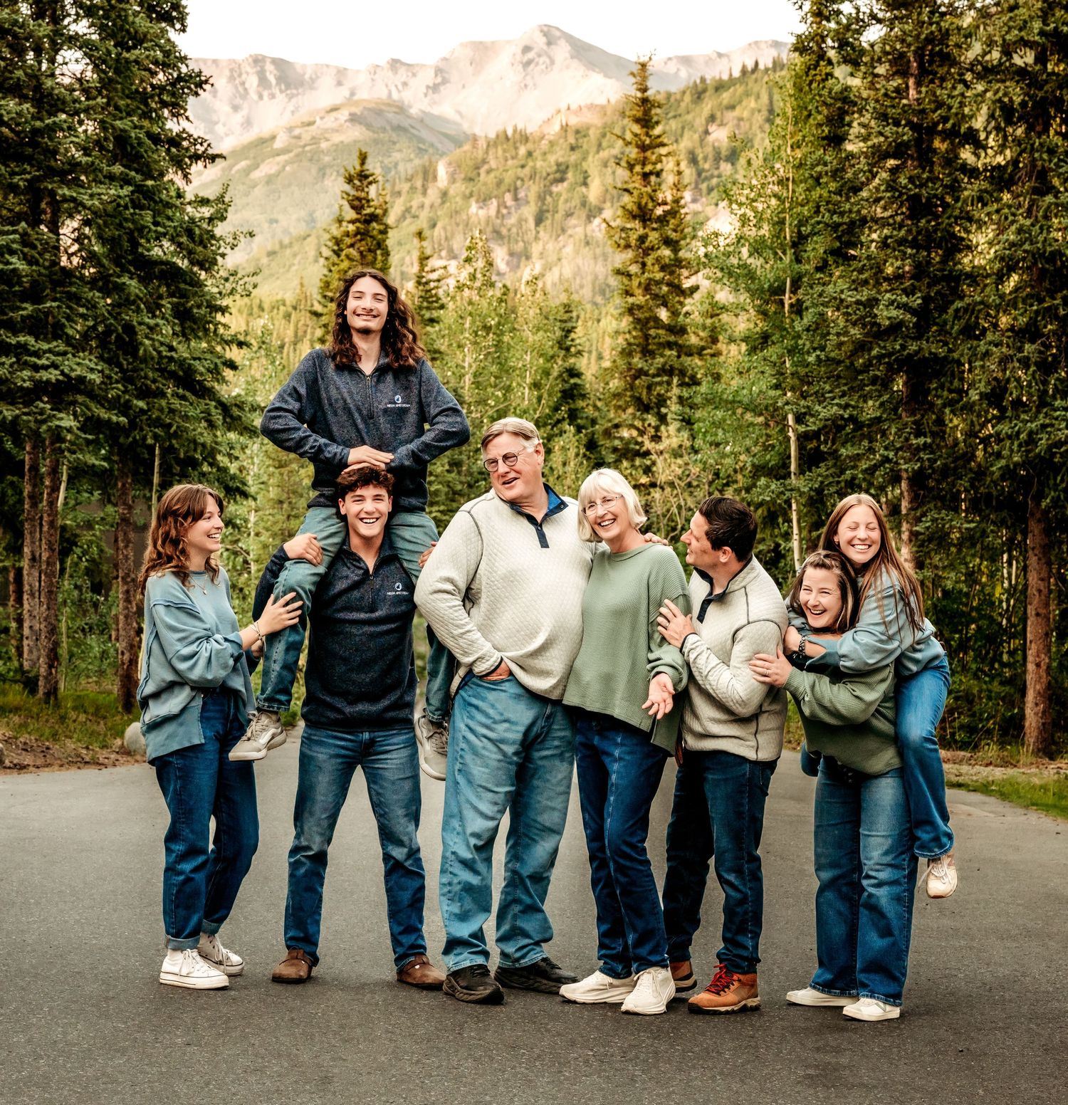A group poses together on a mountain road with evergreen trees and peaks in the background.