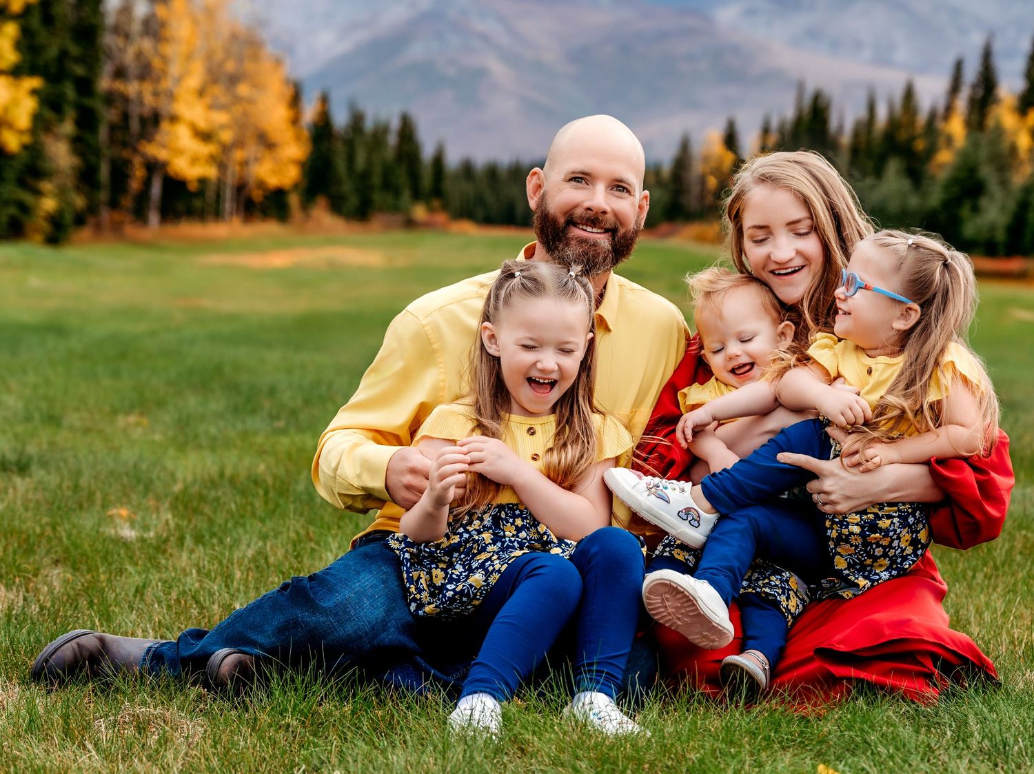 A happy family sits together on grass with mountain and autumn trees in the background.