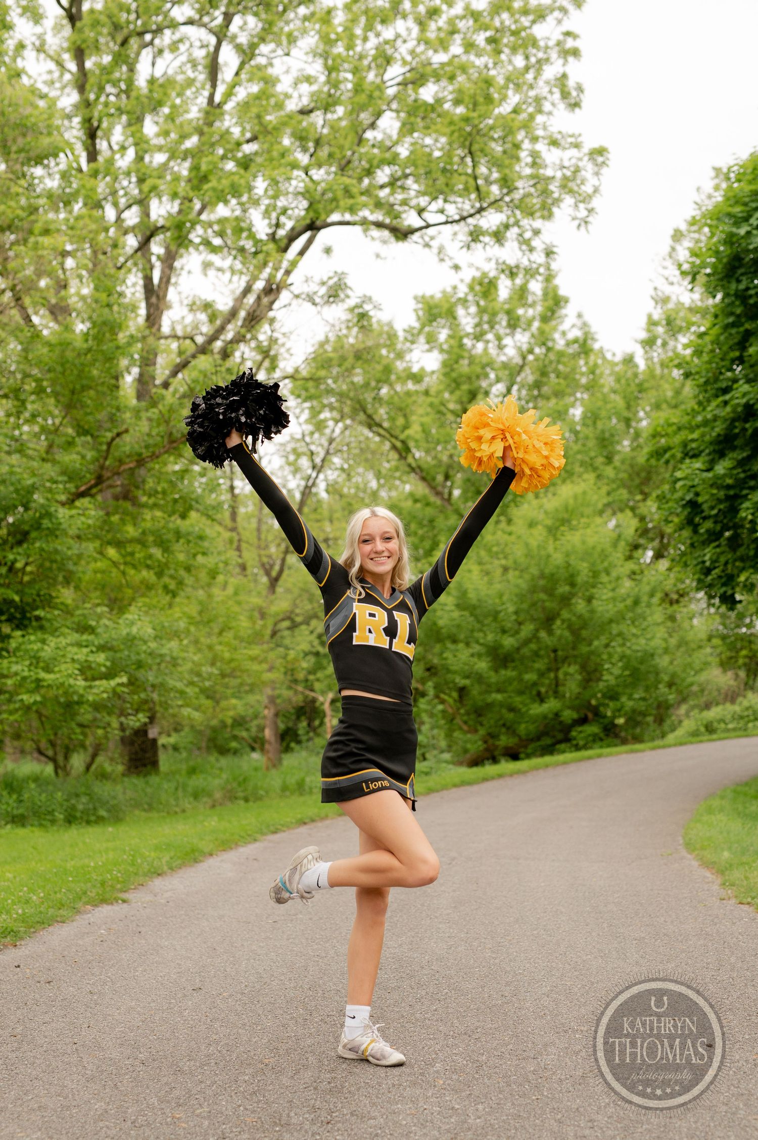 Energetic pose with black and gold pom poms raised high on park pathway surrounded by trees.