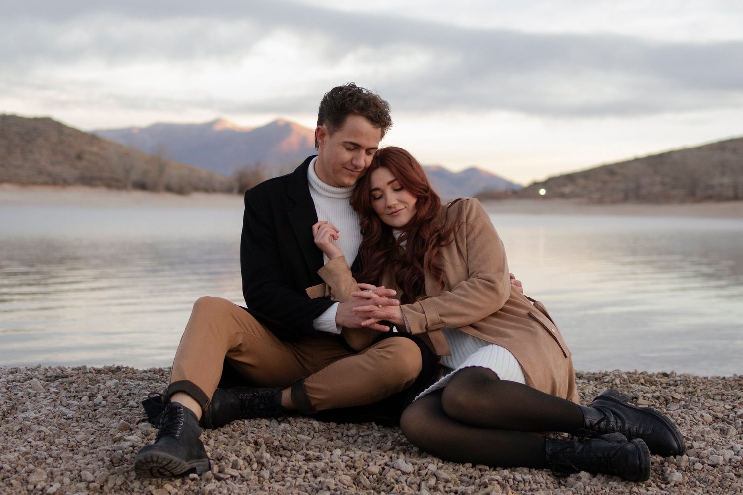 Couple sits on the shoreline, cuddled up and holding hands.