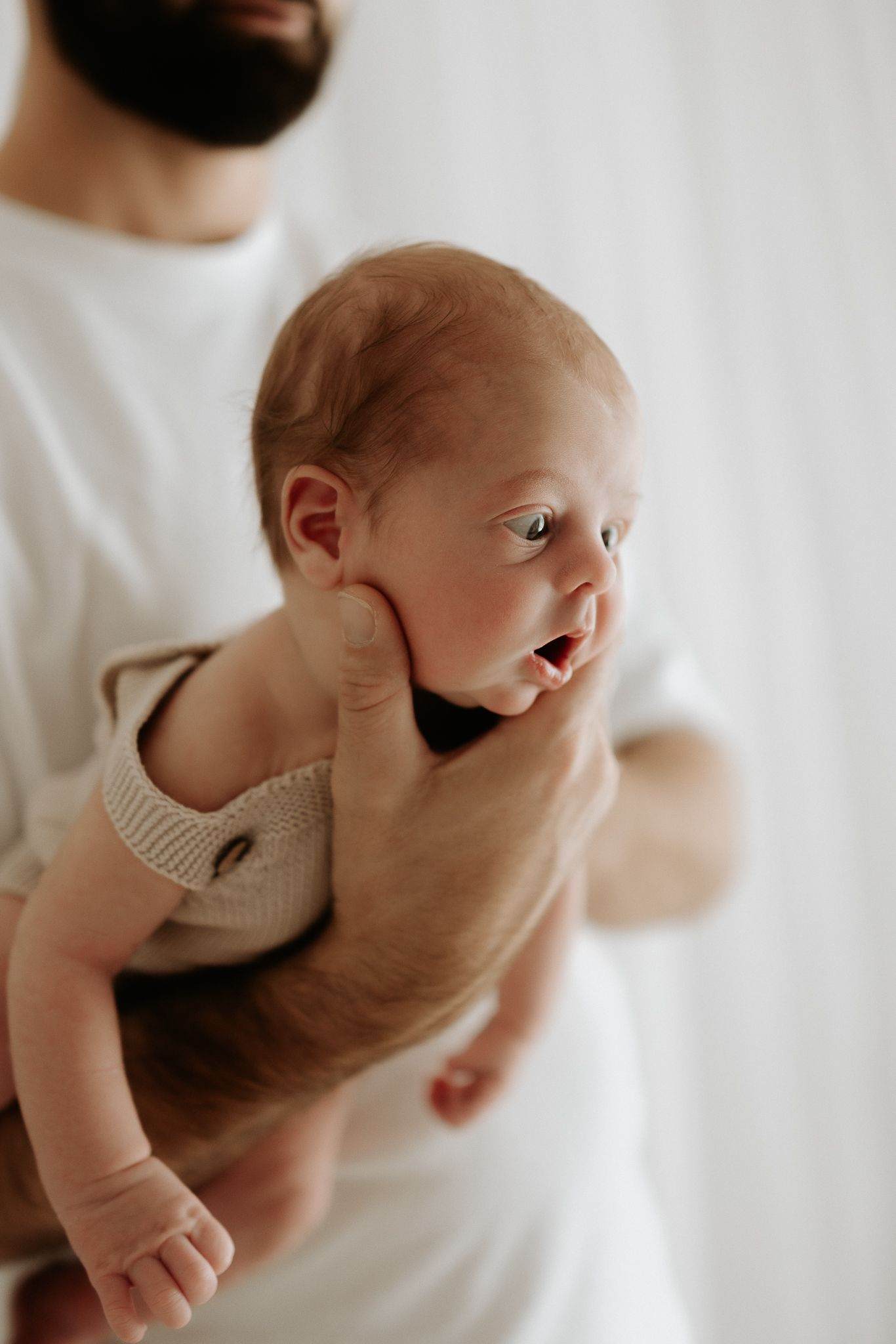 A sleeping newborn baby in white clothing stretches their arms against crisp white bedding.