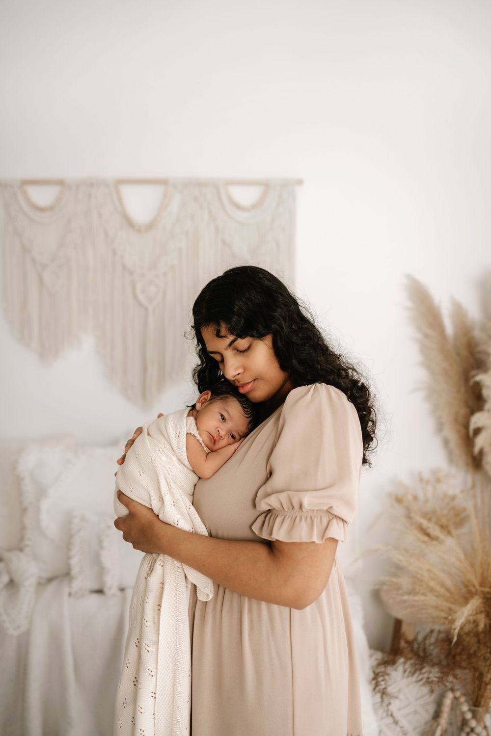 Tender moment between mother and newborn in cream-colored dress against macrame wall hanging.