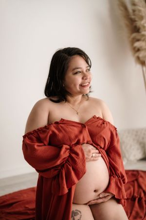 Joyful maternity portrait in rust-colored dress against neutral backdrop with pampas grass.