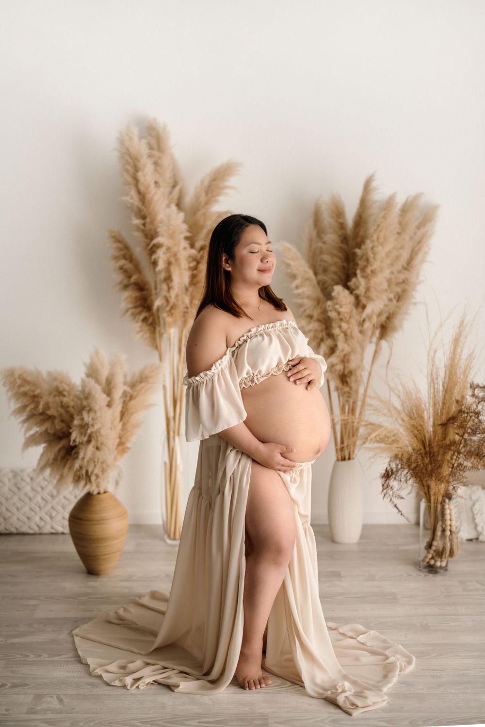 Beautiful maternity portrait with flowing cream dress and pampas grass decor in light minimal studio setting.