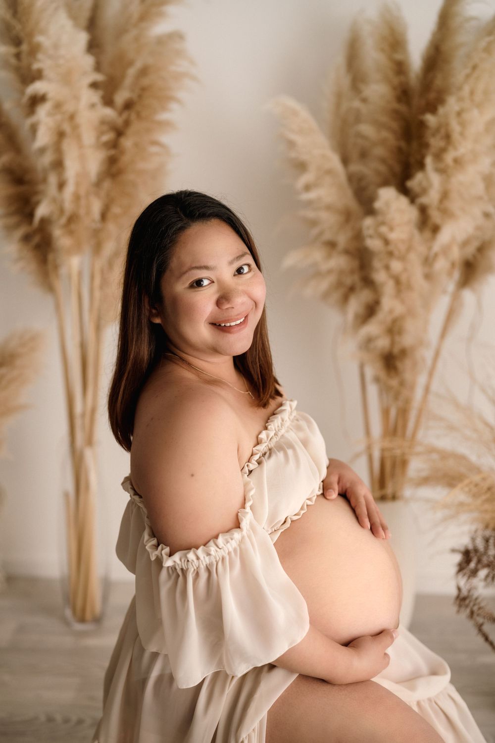 Beautiful maternity portrait with flowing cream dress and pampas grass decor in warm, neutral tones.
