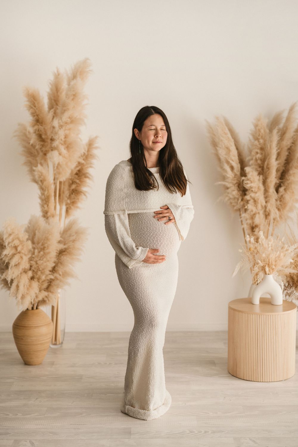Maternity photo series with cream knit dress against neutral background with pampas grass decor in vases.